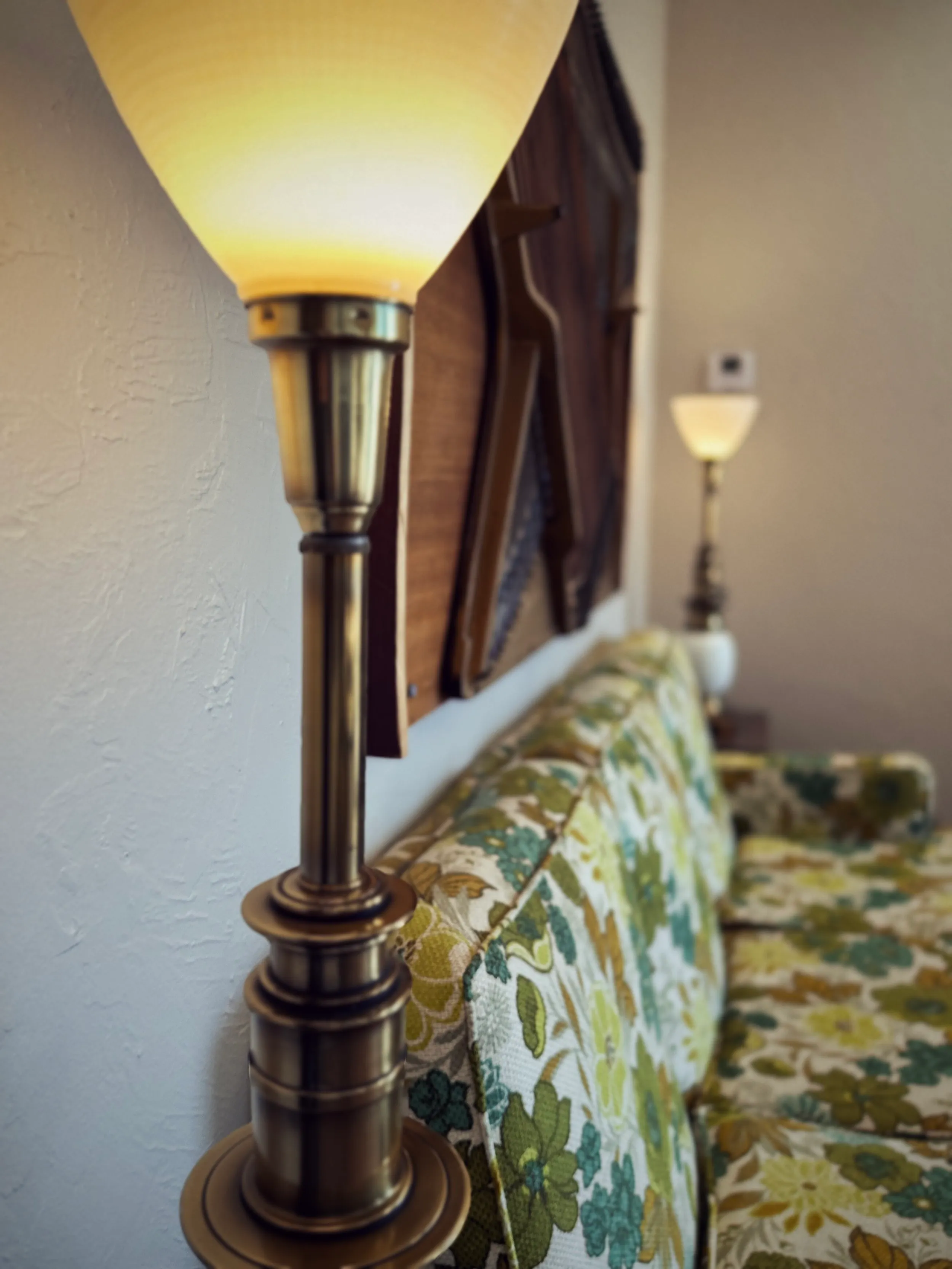 A brass torchiere lamp glowing warmly beside a green floral sofa and a woven upright-piano back