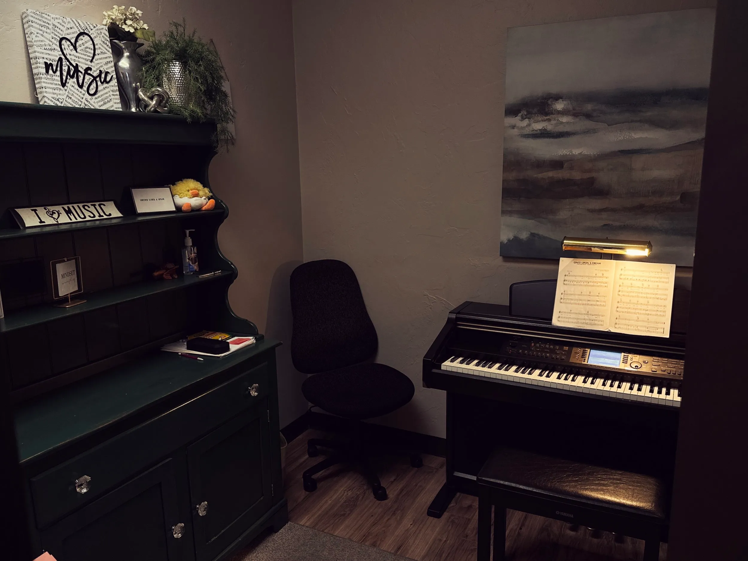 A piano studio with an "I love music" shelf, a digital piano lit by a brass lamp, and sheet music on the stand