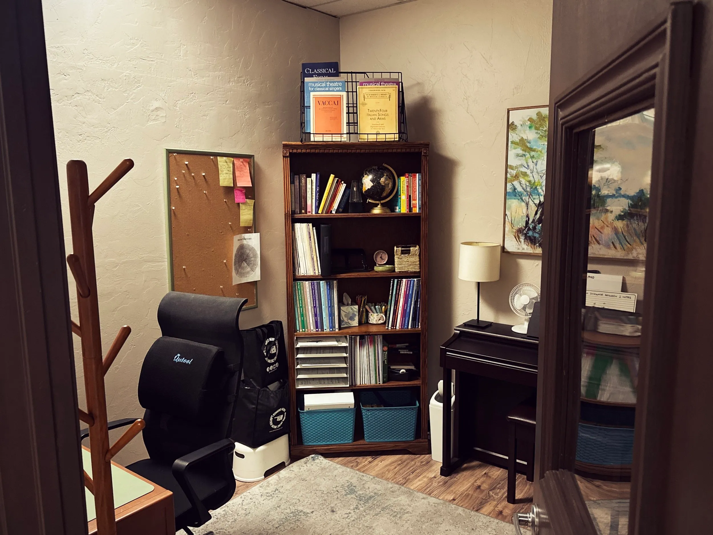 A teaching room with a tall bookshelf of music method books, a cork bulletin board, and a digital piano