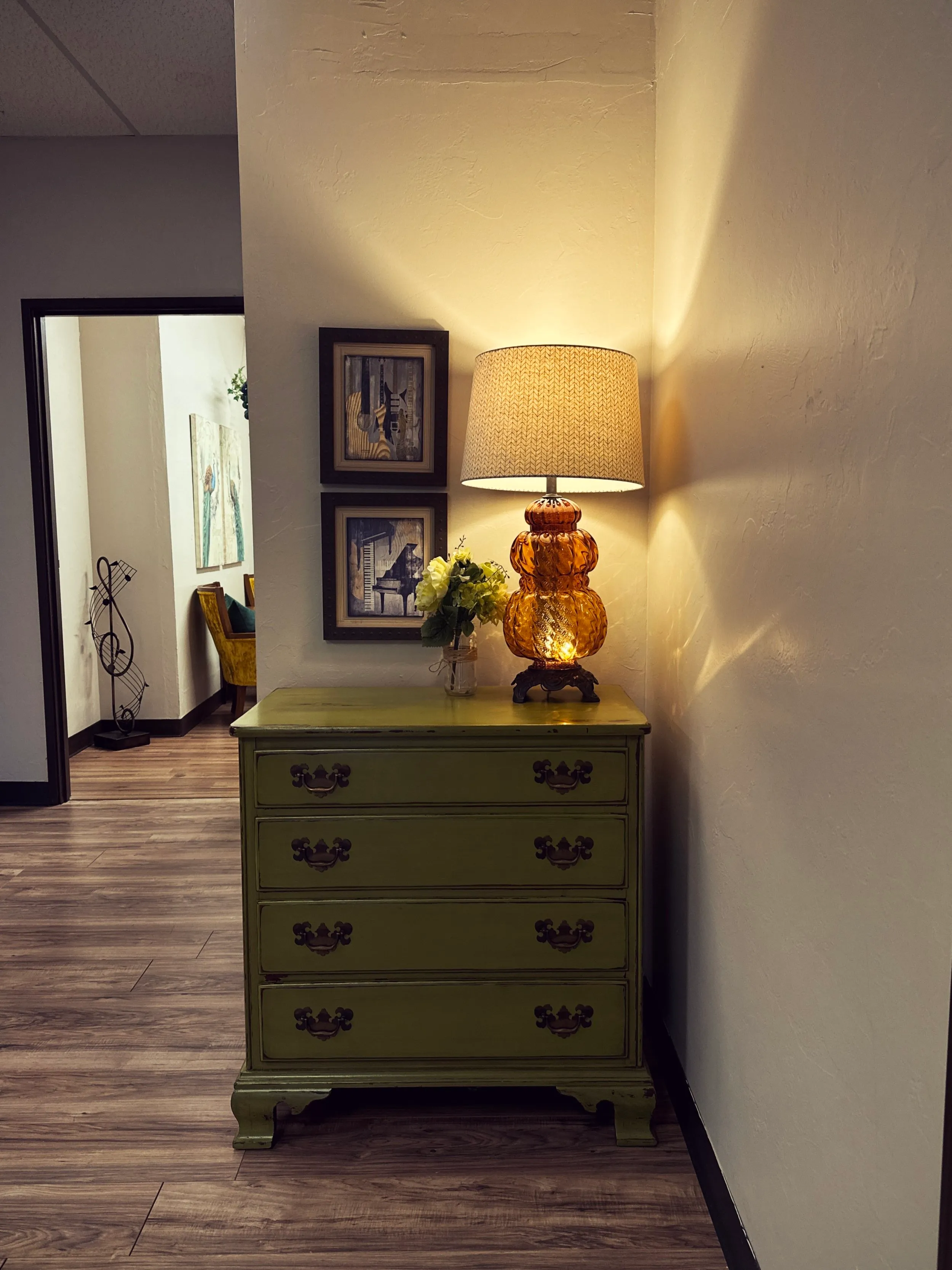 An entry vignette with a sage-green chest, a glowing amber glass lamp, and framed piano prints, looking toward the waiting area